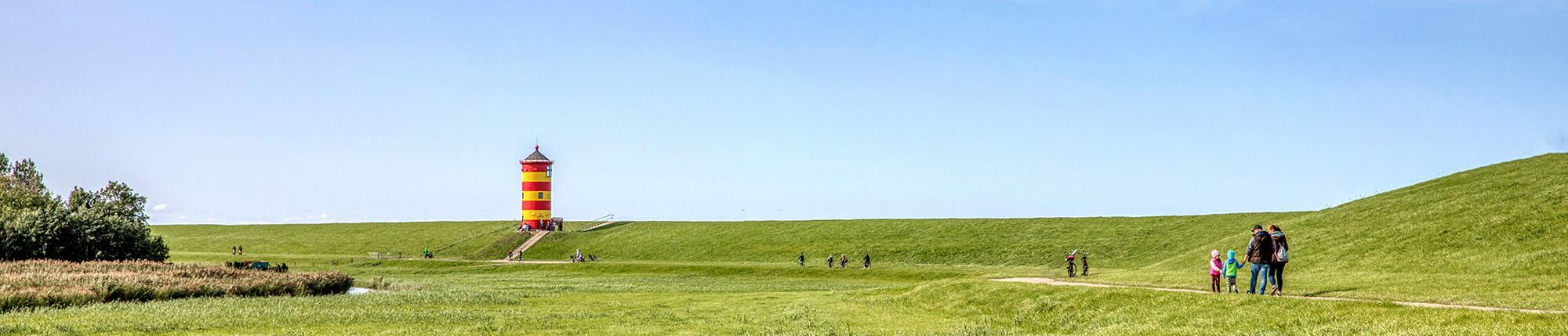Spaziergänger und Radfahrer am Deich mit Leuchtturm in der Ferne